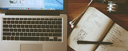 A laptop displays browser tabs; beside it, a notebook holds sketches and text with a pen lying on top. Earbuds and a camera rest nearby on a wooden table.