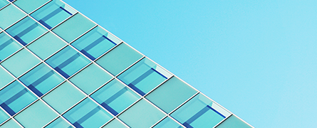 A glass building facade reflects light, with blue-tinted windows arranged in a grid pattern against a clear, cloudless sky.