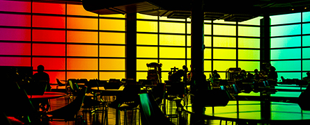 Silhouetted chairs and tables rest in front of a large, colorful gradient window, spanning from red to blue, creating a vibrant backdrop in a modern cafeteria setting.