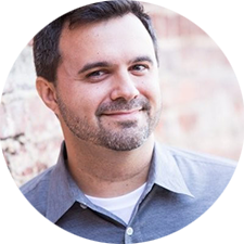 A man with short dark hair and a beard is smiling. He wears a gray collared shirt, standing against a blurred brick wall.