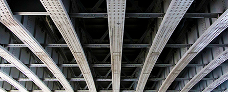 Metal beams span overhead, crisscrossing in a geometric pattern, forming the underside of a bridge. The surrounding area is dark, highlighting the structure's intricate design.