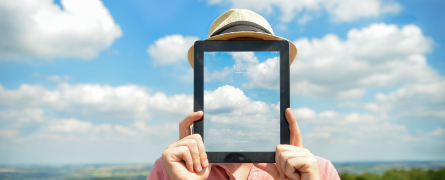 A person holds a tablet in front of their face, displaying a clear sky and clouds that blend with the actual sky background. The person wears a hat, creating a seamless illusion.