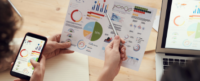 Two hands hold and point to multiple colorful charts on paper, including pie charts and bar graphs, while another hand holds a smartphone displaying similar charts near a laptop on a wooden table.