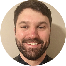 A person with dark hair and a beard is smiling, facing the camera, wearing a dark jacket and a dotted shirt. The background is a plain, light-colored wall.