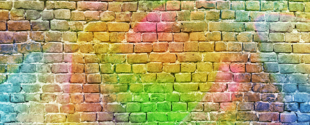 A brick wall displays vibrant, multicolored spray paint patterns, blending hues of red, green, blue, and yellow, creating an abstract artistic effect. The setting is urban and visually striking.