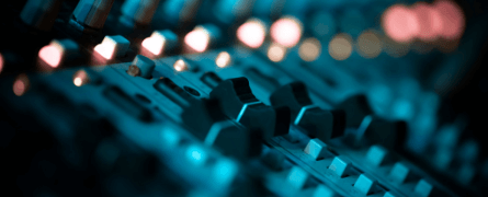 Audio mixer with multiple knobs and sliders, illuminated by soft blue and pink lighting, set in a dimly lit studio environment.