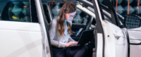 A woman uses a laptop while seated in the open driver's door of a white car, surrounded by a modern, industrial-style indoor environment. No text is present.