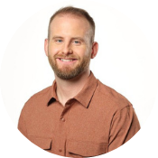 A man wearing a brown shirt smiles gently, standing against a plain white background.