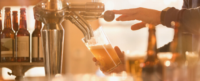 A hand pours beer from a tap into a glass, surrounded by various bottles on a bar counter, with warm lighting enhancing the scene.