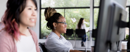 Two people work at computers, smiling and focused. They're in a bright office with large windows showing a green, leafy view outside.