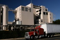 A red semi-truck drives past a large industrial building with multiple levels and storage silos, set against a clear blue sky.