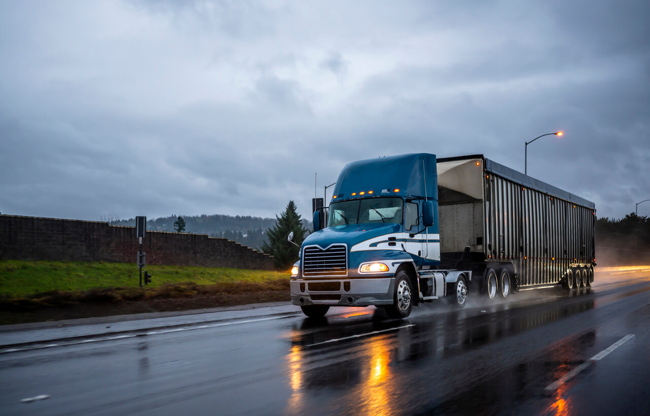 A large blue semi-truck travels on a wet highway, emitting spray from the road. It is surrounded by overcast skies and distant trees, with streetlights lining the roadside.