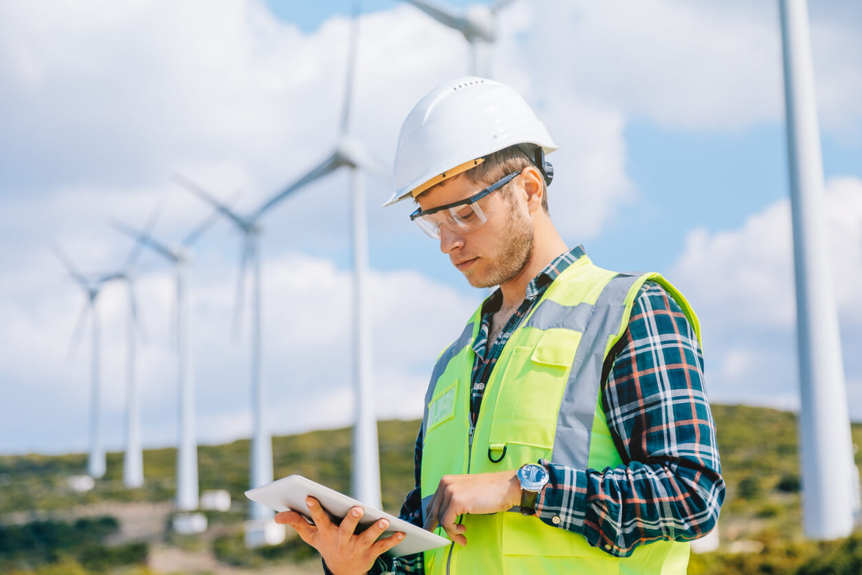 A person wearing a hard hat and safety vest uses a tablet in front of several wind turbines on a sunny day, set in a rural landscape.