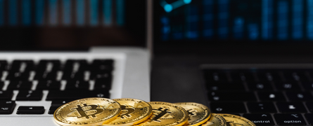 Golden Bitcoin coins rest on a laptop keyboard, positioned between two laptops displaying blue digital graphs in a dimly lit setting.