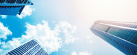 Skyscrapers stretch skyward, reflecting sunlight against a backdrop of bright blue sky and scattered white clouds. The scene captures a modern urban environment from a low-angle perspective.