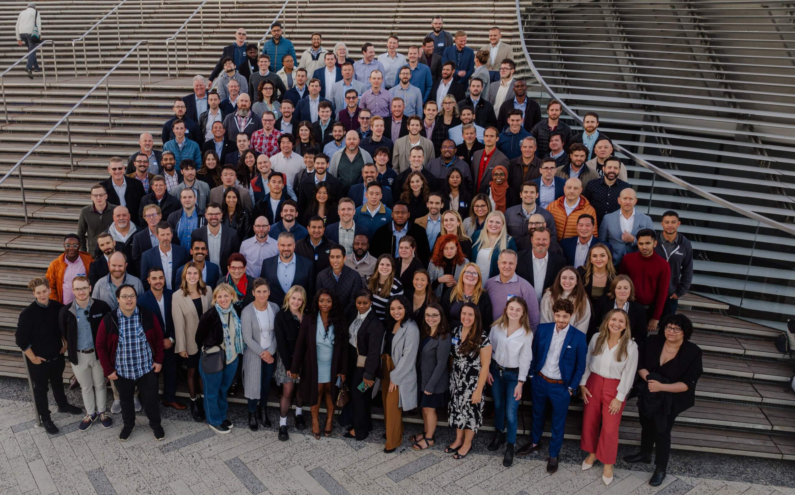 A large group of diverse people, standing closely together, are posing on a wide, tiered staircase in an outdoor setting. Everyone is smiling, suggesting a happy or celebratory occasion.