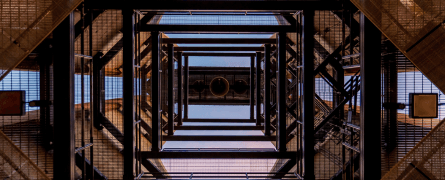 Metallic stairwell spirals upward, viewed from below, creating geometric patterns. The structure's grid-like framework is contrasted against a clear blue sky, emphasizing architectural symmetry.