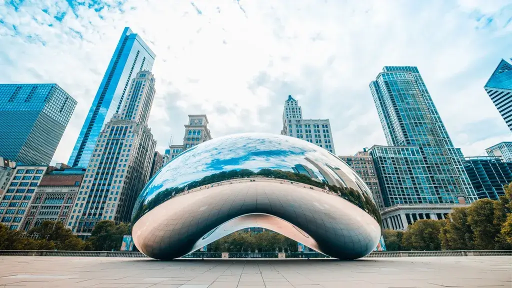A large, reflective bean-shaped sculpture sits amid a plaza, surrounded by towering skyscrapers and a cloudy sky.
