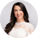A person with long, dark hair smiles warmly while wearing a white lace blouse, set against a plain, light gray background.