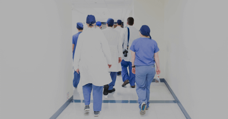 A group of healthcare professionals in scrubs and lab coats walking down a hospital hallway, viewed from behind.