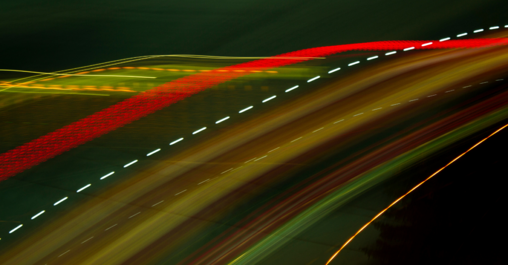 Colorful light trails streak across a dark road at night, forming curved, dynamic lines.