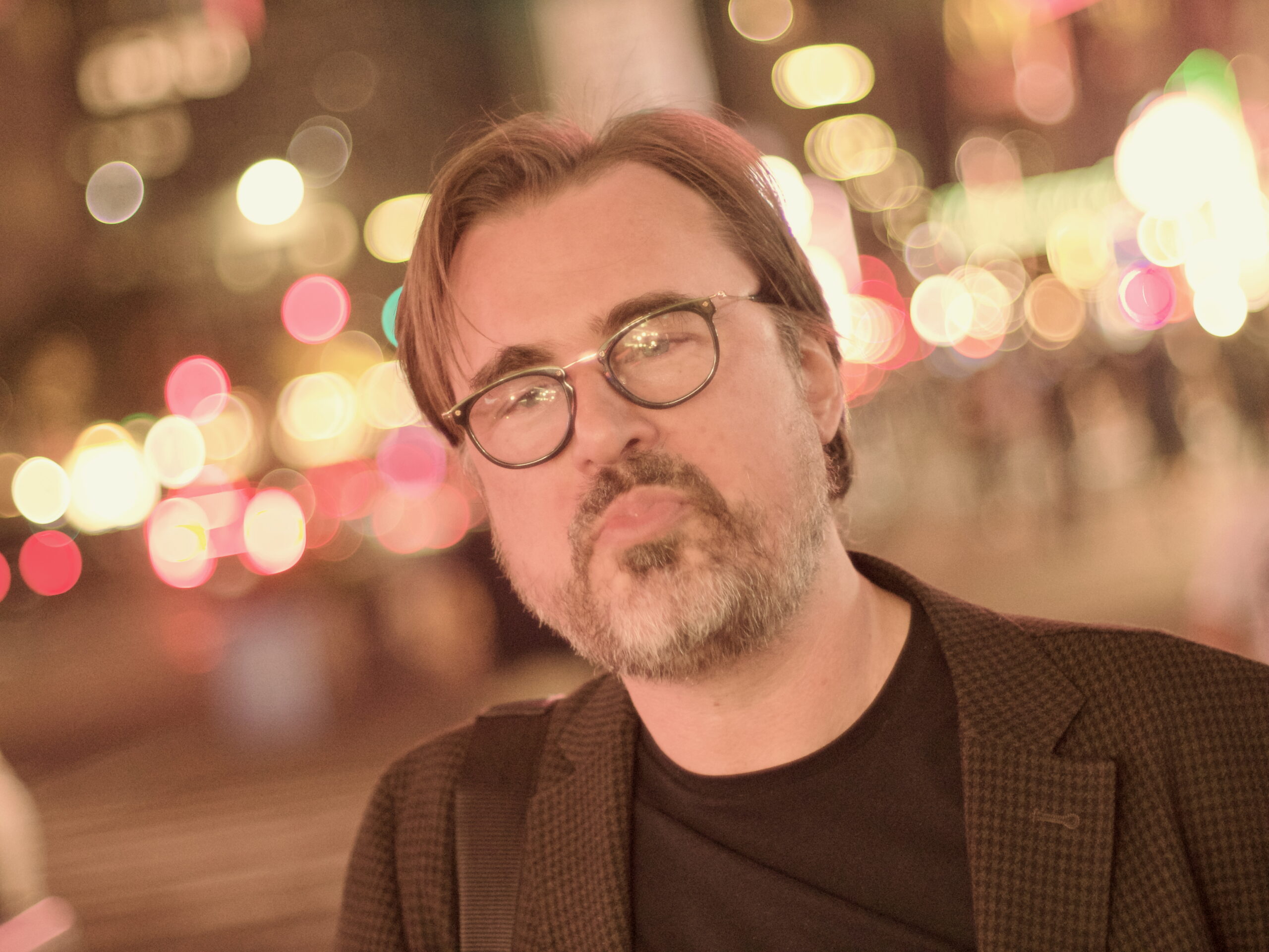 A man with glasses and a beard wearing a dark blazer stands on a city street at night with blurred colorful lights in the background.